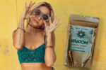 woman with blonde hair wearing blue green tube top posing and holding her sunglasses beside a kratom powder package