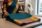 A person’s hands holding a Semi Kanchipuram silk saree with gold zari border on a boutique display table, with clothing racks blurred in the background.